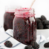 jar of blackberry freezer jam with a spoon in it. white napkin with blue plaid underneath. fresh blackberries scattered around