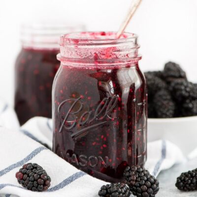 jar of blackberry freezer jam with a spoon in it. white napkin with blue plaid underneath. fresh blackberries scattered around