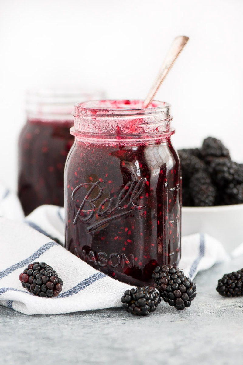 jar of blackberry freezer jam with a spoon in it. white napkin with blue plaid underneath. fresh blackberries scattered around