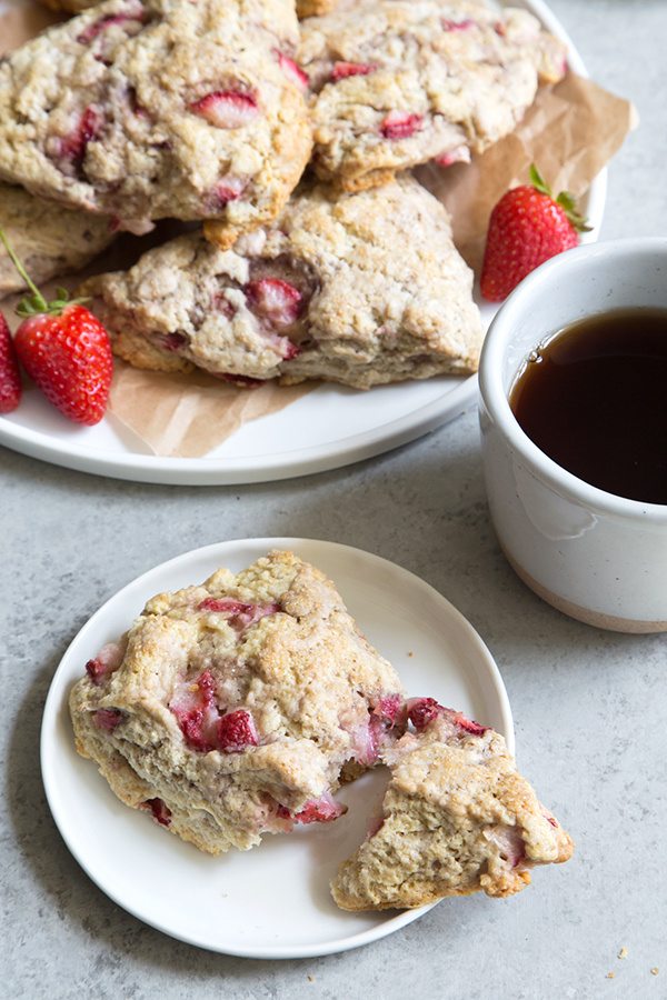 Strawberry Buttermilk Scone on a white plate broken apart to eat. Cup of coffee and platter of more scones in the background with fresh strawberries