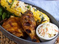 carolina style barbecue chicken in a serving dish with cole slaw and corn on the cob on a woven mat with a blue/white striped napkin