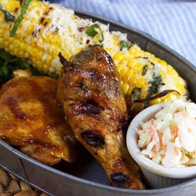 carolina style barbecue chicken in a serving dish with cole slaw and corn on the cob on a woven mat with a blue/white striped napkin