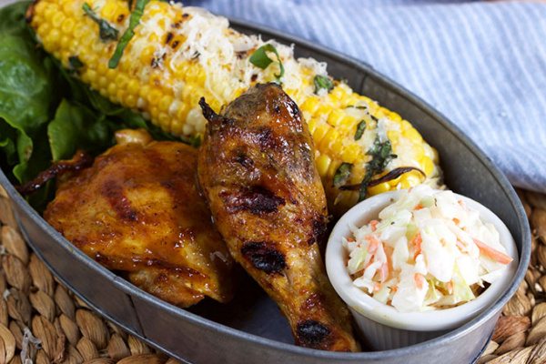 Carolina Style Barbecue Chicken in a metal serving dish with corn on the cob and cole slaw on a woven mat with a white/blue striped napkin on the side