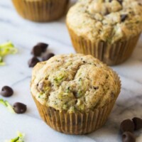 zucchini chocolate chip muffins sitting on a marble surface with chocolate chips and shredded zucchini scattered about