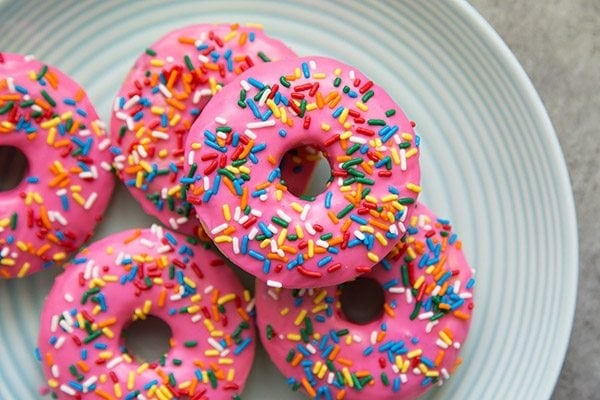 a white plate of pink glazed doughnuts with rainbow sprinkles