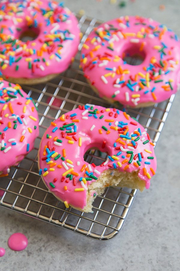 Birthday Cake Pink Glazed Doughnuts with rainbow sprinkles on a rack one with a bite taken out