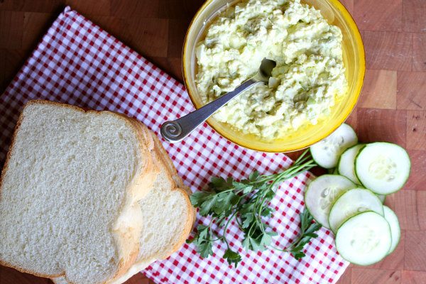 Yellow bowl of Avocado Egg Salad with a spoon in it and slices of bread, cucumber and parsley on the side. set on a white/red checked napkin