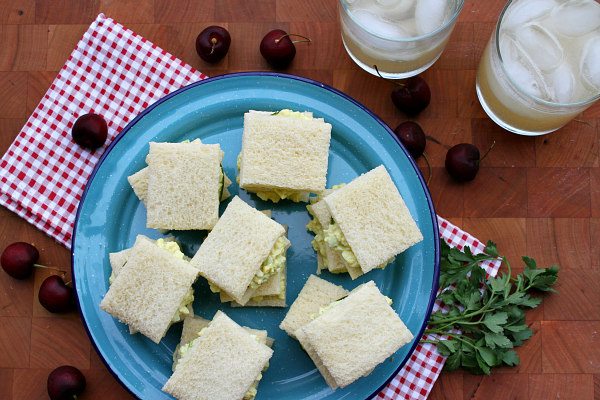 overhead shot of Avocado Egg Salad Tea Sandwiches on a blue plate set on a red/white checked napkin with cherries scattered around and two glasses of lemonade