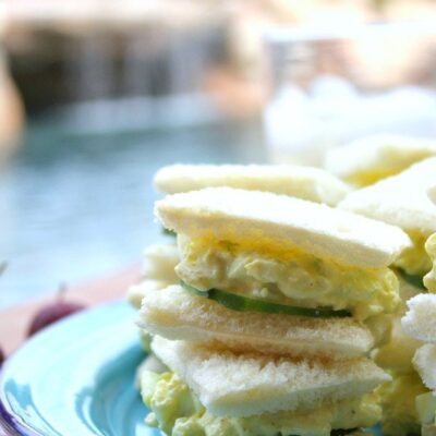 avocado egg salad tea sandwiches stacked on a blue plate on a red/white checked tablecloth with pool backyard setting