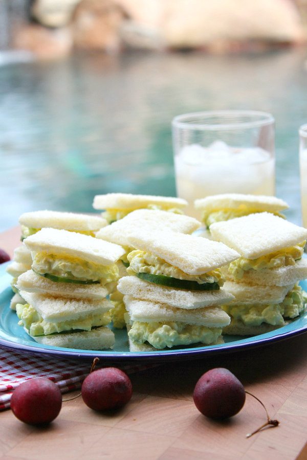 Avocado Egg Salad Tea Sandwiches stacked on a blue plate with fresh cherries scattered around and glass of lemonade in the background of a backyard pool setting