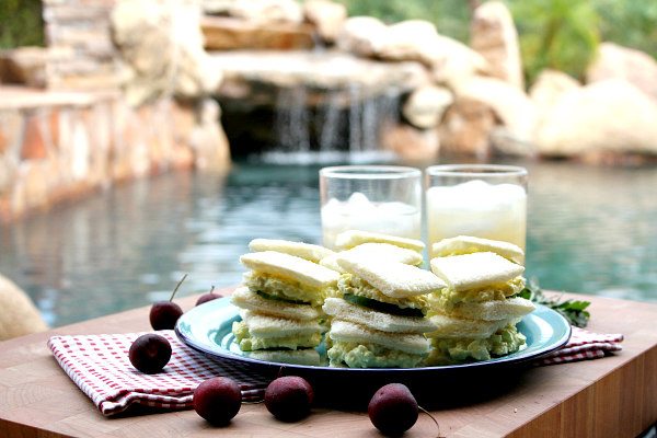Avocado Egg Salad Tea Sandwiches stacked on a blue plate set on top of a red/white checked napkin on a cutting board. two glasses of lemonade in the background. fresh cherries scattered around. pool backyard setting