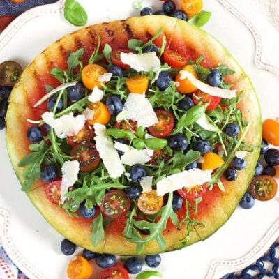 Grilled Watermelon Pizza with Blueberries, Parmesan and Arugula on a white plate with more tomatoes and fruit and a wedge of parmesan in the background