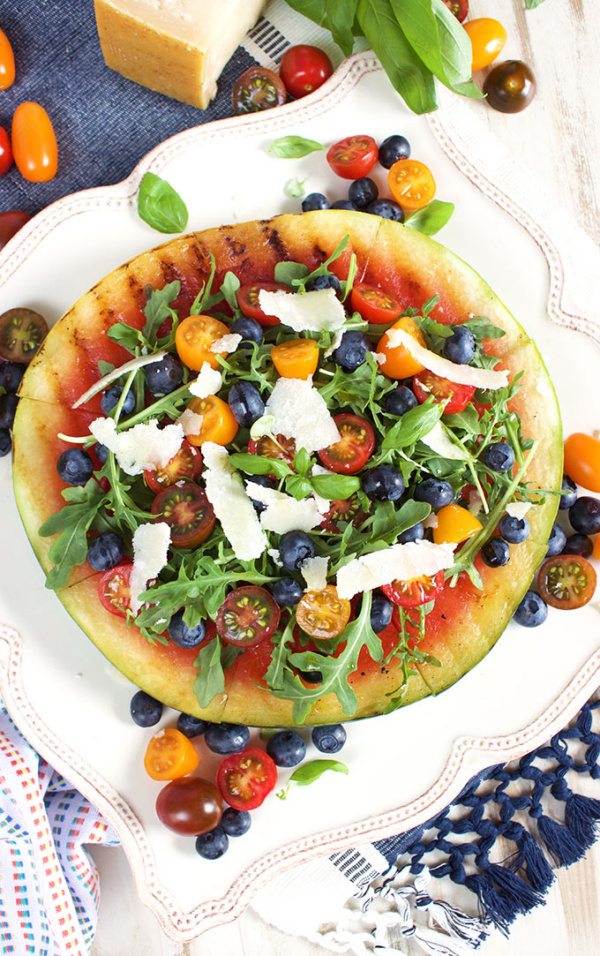 Grilled Watermelon Pizza with Blueberries, Parmesan and Arugula on a white plate with additional tomatoes and fruit on the plate and in the background set on various color blue and patterned napkins. wedge of parmesan in background