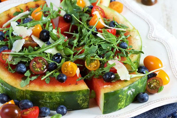 Slicing a wedge of Grilled Watermelon Pizza with Blueberries, Parmesan and Arugula displayed on a white plate