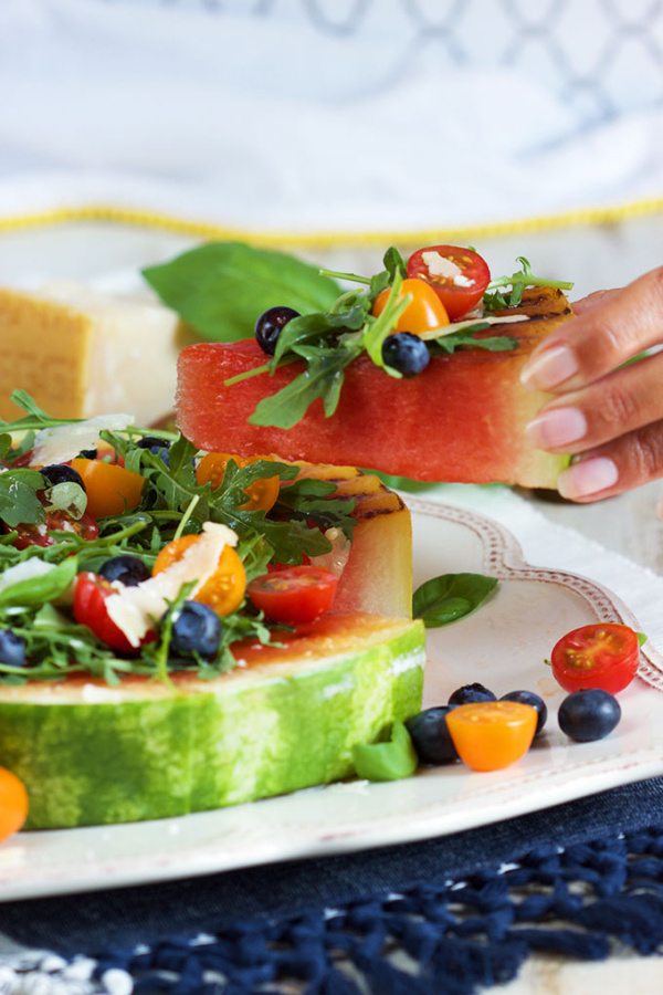 Hand holding Slice of Grilled Watermelon Pizza with Blueberries, Parmesan and Arugula displayed on a white plate on top of a blue placemat