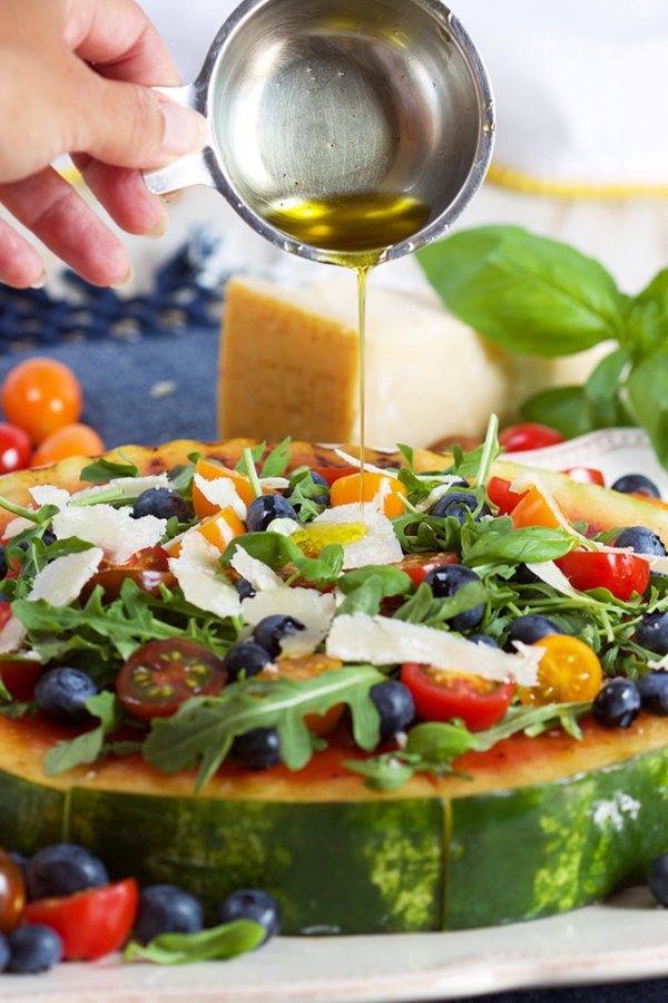 hand Adding oil to Grilled Watermelon Pizza with Blueberries, Parmesan and Arugula with wedge of parmesan cheese in the background