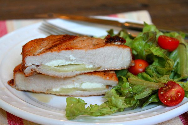 Blue Cheese and Apple Stuffed Grilled Pork Chops cut open to see the inside, displayed on a white plate with a fork and knife and lettuce/tomato salad