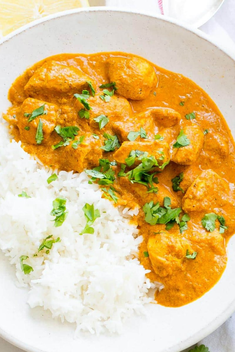 overhead shot of single serving of indian butter chicken in a white bowl served with white rice and cilantro garnish