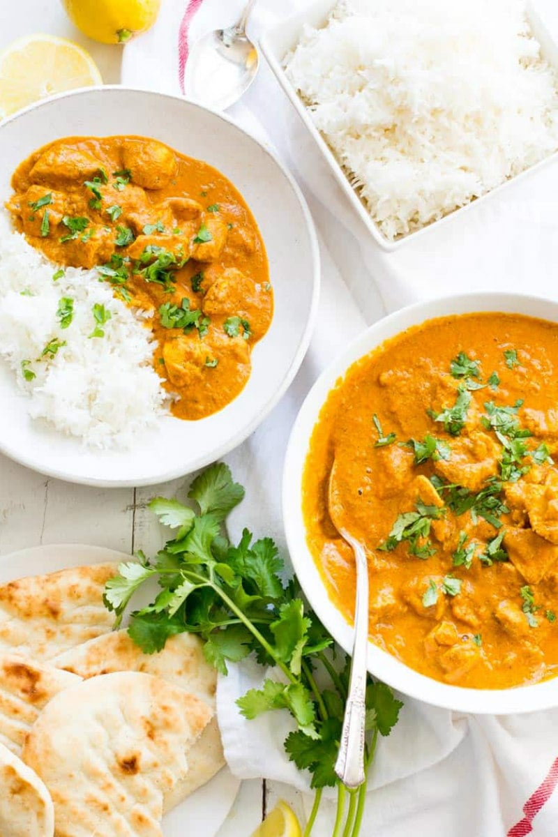 overhead shot of two servings of indian butter chicken in white bowls with white rice and cilantro garnish and pita bread on the side