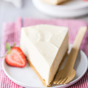 No-Bake Cheesecake slice on a white plate with a fork and half strawberry sitting on a pink/white checked napkin. another slice in view in the background