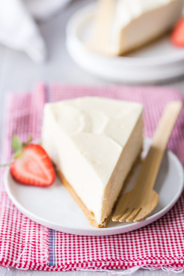 slice of no bake cheesecake on a white plate with fork and strawberry, sitting on a red/white checked napkin. peek at another slice of cheesecake in the background