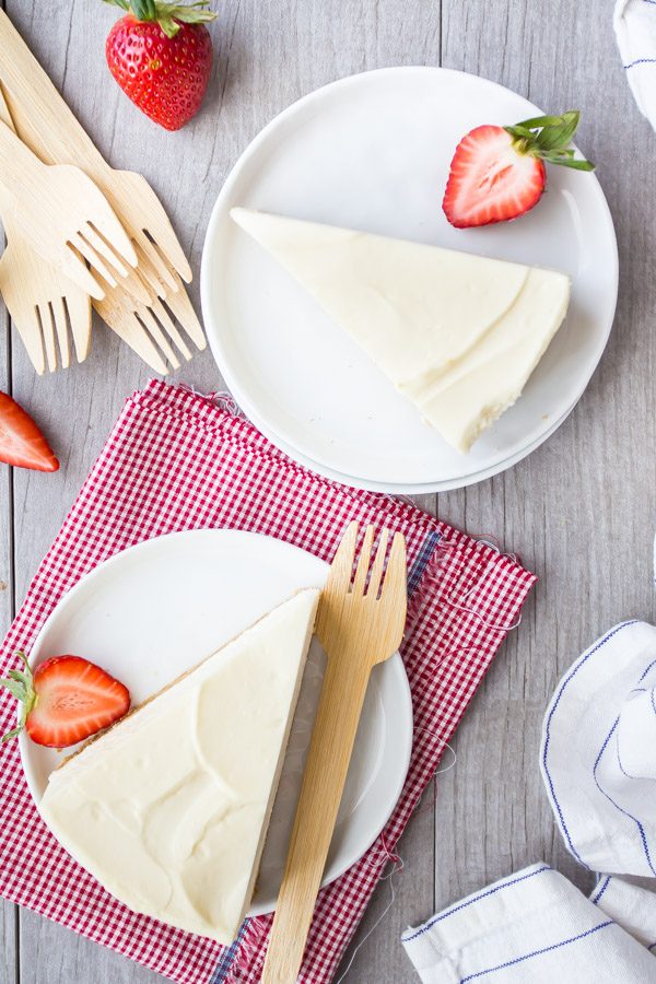 overhead shot of slices of no bake cheesecake on white plates with forks and strawberries. white/red checked napkin and blue/white striped napkin in background