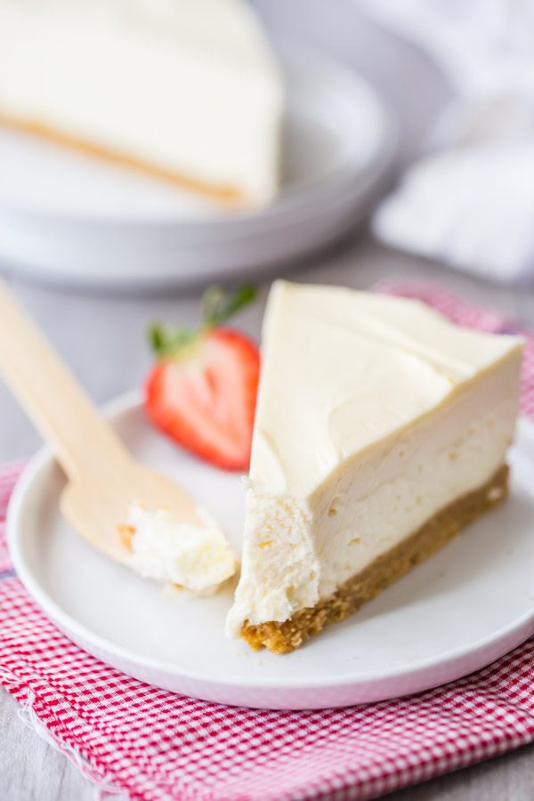 slice of no bake cheesecake with a bite out of it on a white plate with a wooden spoon and a strawberry. sitting on a white and pink checked napkin