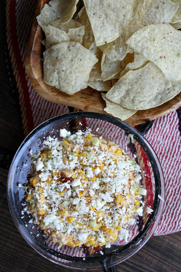 Slow Cooker Mexican Street Corn Dip in a glass bowl with bowl of Tortilla Chips set on a colorful Mexican blanket