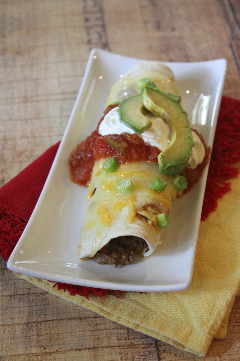 single serving of beef burrito casserole with sour cream, salsa and avocado on a white plate with yellow and red napkins underneath