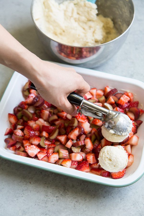 Adding Biscuits to make Strawberry Rhubarb Cobbler in a white casserole dish. bowl of biscuit batter in the background