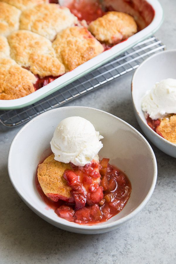 Strawberry Rhubarb Cobbler served with ice cream in a white bowl with the pan of cobbler in the background sitting on a cooling rack