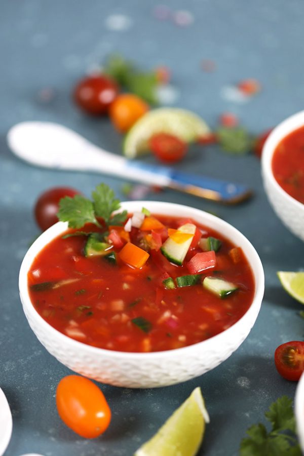 white Bowl of Gazpacho garnished with tomato, cilantro and lime. Spoon in the background.