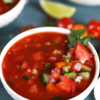 gazpacho in a white bowl garnished with tomato and cilantro. lime wedge in background and peek at two more bowls of gazpacho
