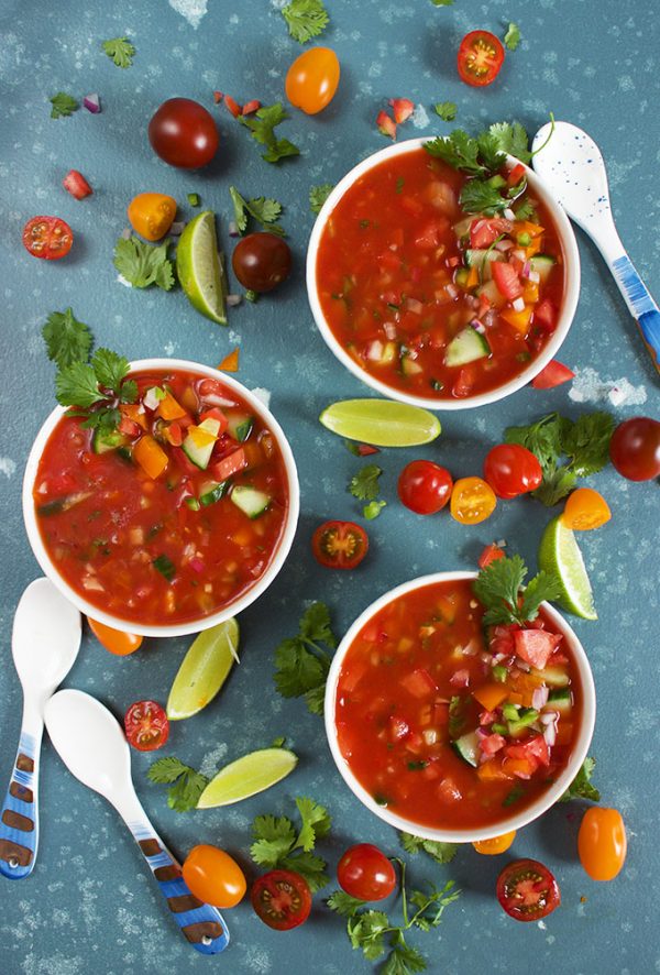 Overhead shot of three Bowls of freshly made Gazpacho garnished with tomato, cilantro and lime. spoons in view too.