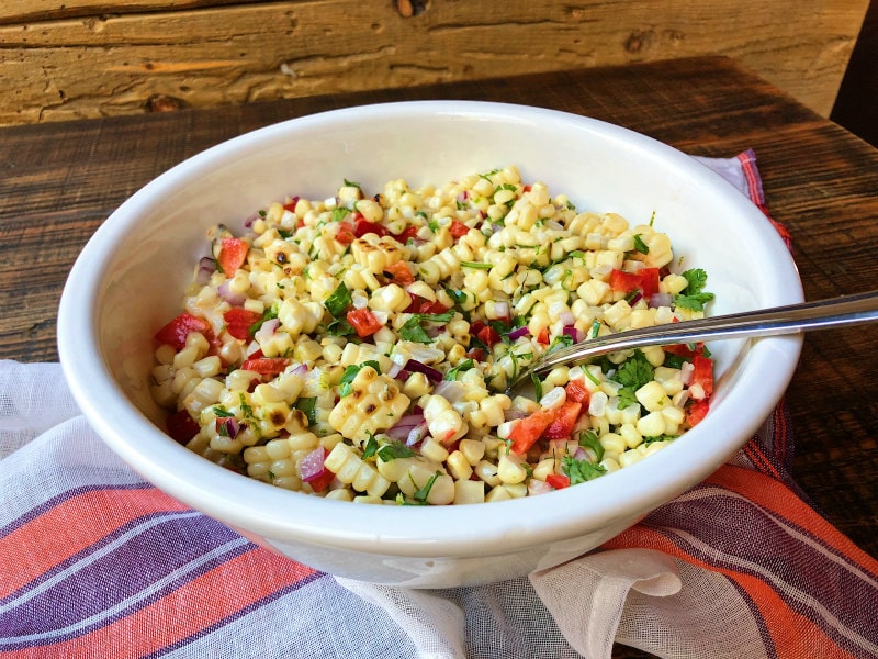 Grilled Corn Salad in a white bowl with a serving spoon sitting on a wooden surface with an orange, purple and white striped cloth napkin