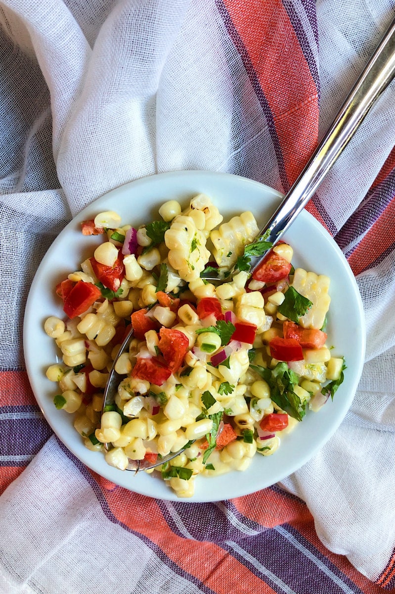 Grilled Corn Salad served in a white bowl with a spoon, Sitting on an orange, purple and white striped cloth napkin