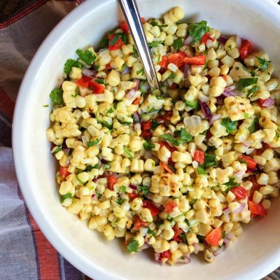 grilled corn salad in a white bowl with a serving spoon on top of an orange, purple and white striped cloth napkin