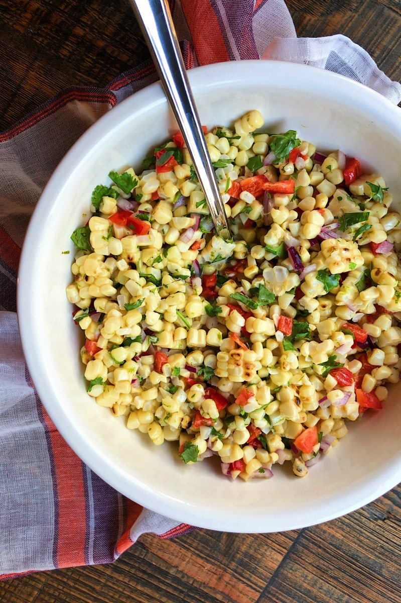 Grilled Corn Salad in a white bowl with a serving spoon set on a wooden board with an orange, purple and white striped cloth napkin