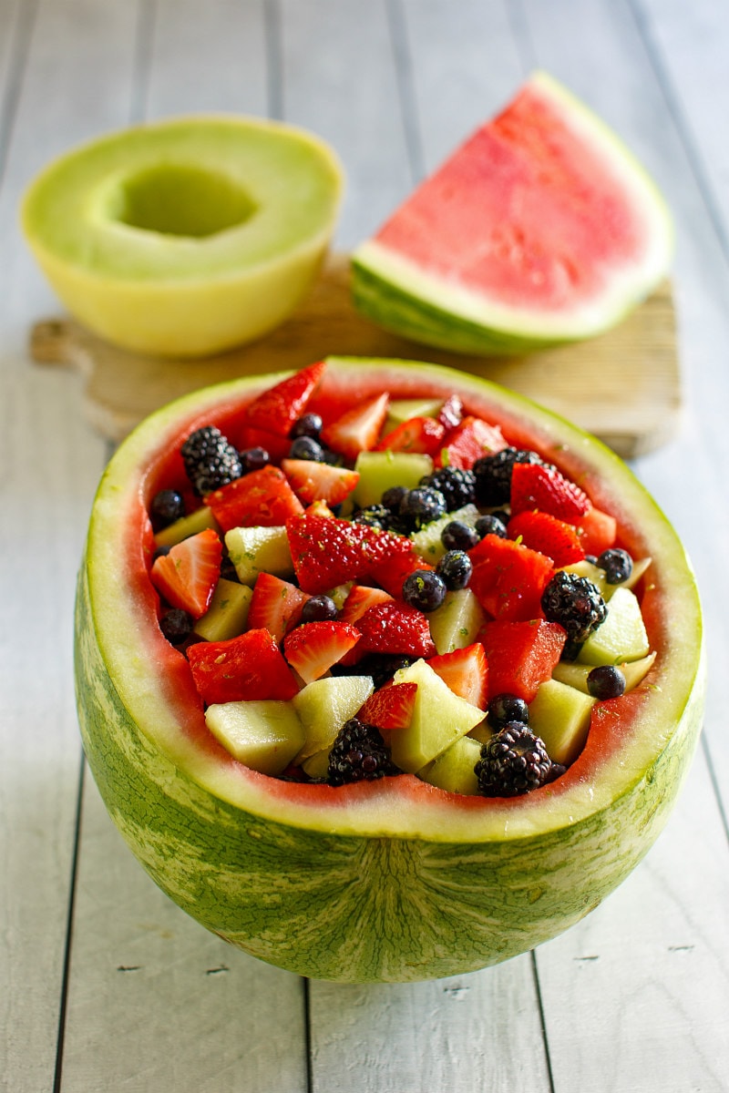 half of a watermelon hollowed out and filled with spiked fruit salad. melon and watermelon sitting on a cutting board in the background