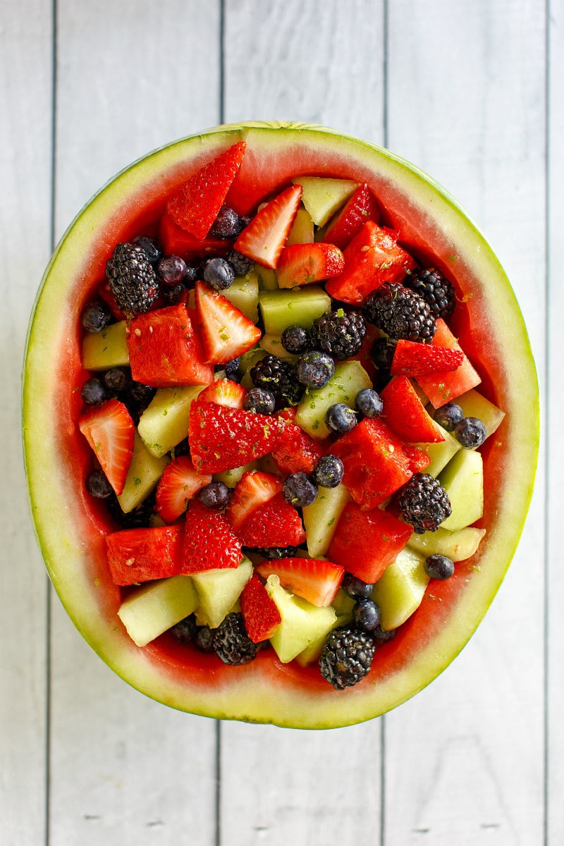 overhead shot of half a watermelon hollowed out and filled with spiked fruit salad. sitting on a white wood plank board surface