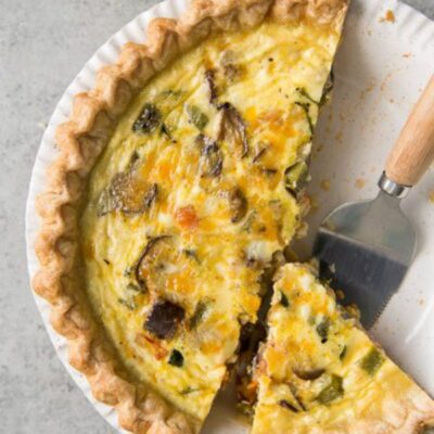 Summer Vegetable Quiche in a white pie plate with a couple of pieces cut out and a pie server on the plate