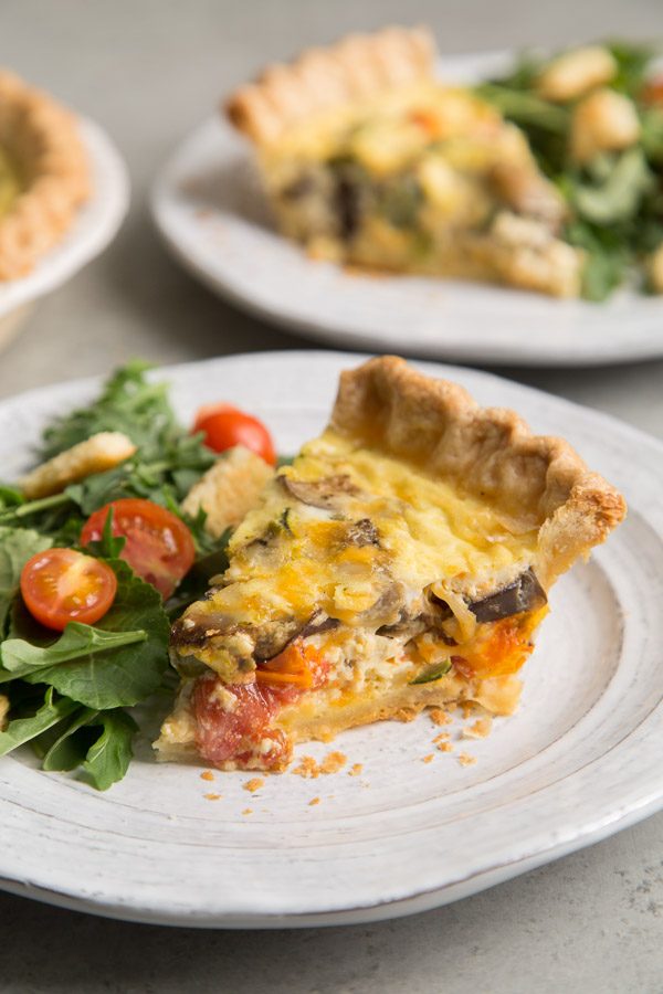 Slice of Summer Vegetable Quiche on a white plate with salad and more plated quiche in the background