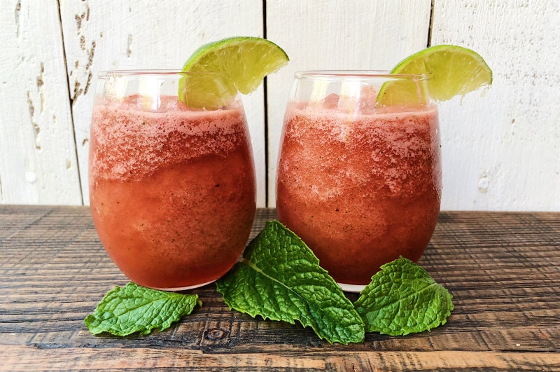 Two Slushie Watermelon Mojitos set on a dark wood table with a white wood backdrop. Glasses garnished with lime wedges and mint leaves in front of glasses