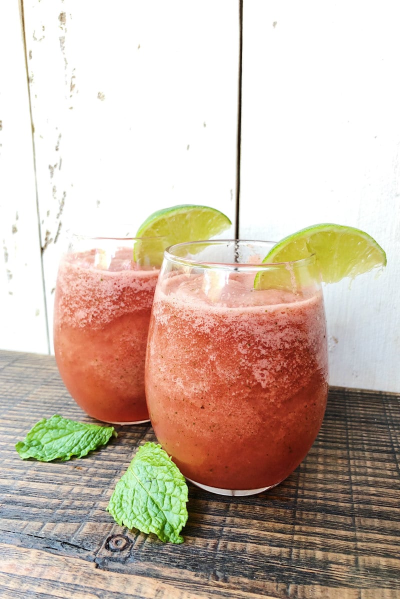 Two Slushie Watermelon Mojitos in glasses on a dark wood table with a white wood Back Drop. Garnished with lime wedges and fresh mint leaves on the table