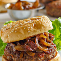 Bacon Burger with Balsamic Caramelized Onions on a white serving platter with lettuce leaf. More buns, burgers and bowl of caramelized onions in the background.