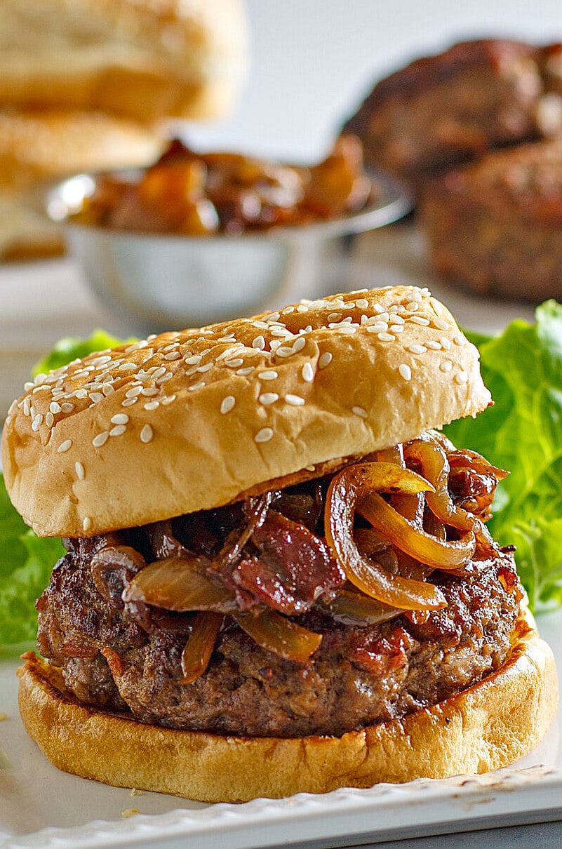 Bacon Burgers with Balsamic Caramelized Onions on a white platter with lettuce in the background and a bowl of caramelized onions in the background