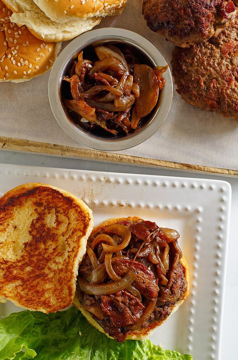 Bacon Burgers with Balsamic Caramelized Onions displayed open faced on a white platter with a lettuce leaf. Bowl of caramelized onions on the side with more buns and burgers