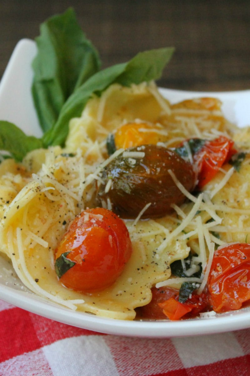 brown butter ravioli with tomatoes garnished with fresh basil on a white plate set on a red/white checked napkin