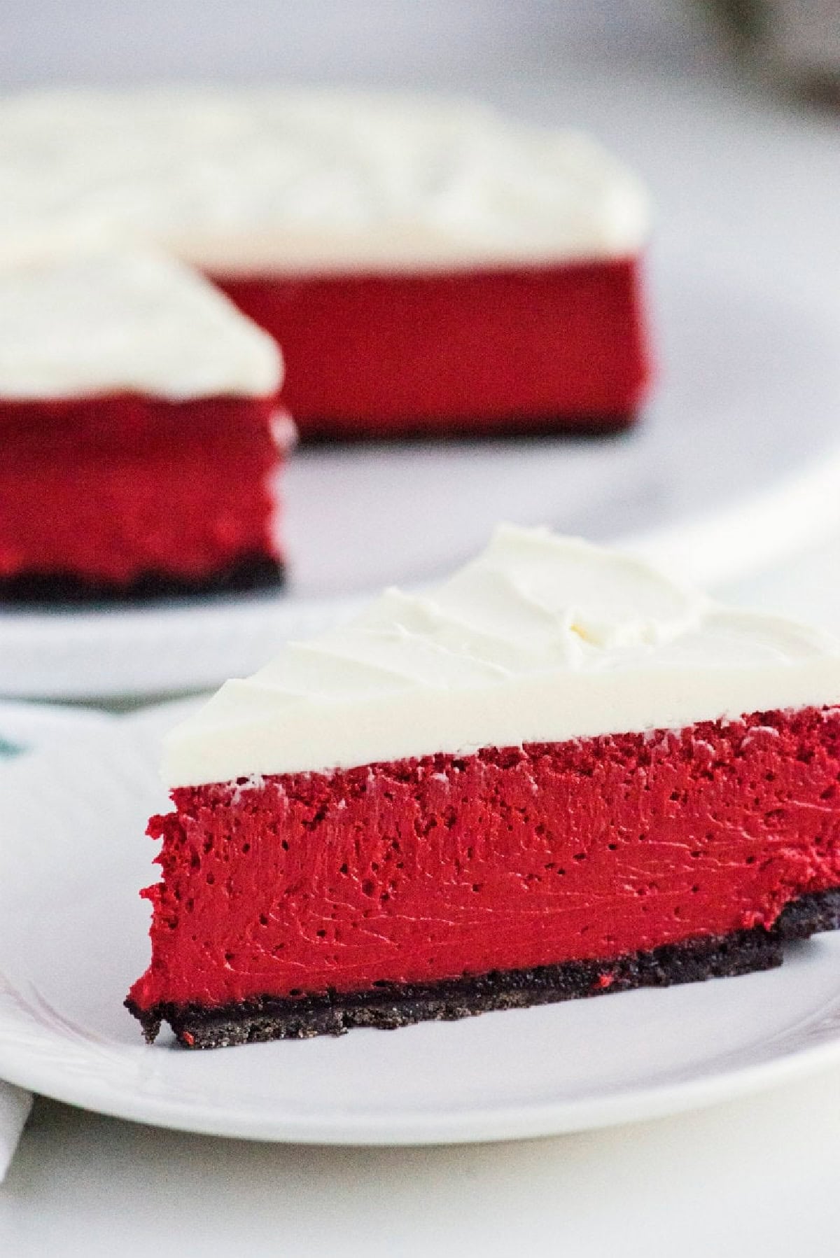 This gorgeous Red Velvet Cheesecake has a creamy red velvet filling, chocolate Oreo crust and rich cream cheese frosting. It's an impressive make-ahead dessert for Christmas, Valentine's Day, holidays and special occasions. slice of red velvet cheesecake with rest of cheesecake in the background
