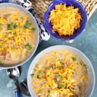 Overhead shot of two bowls of creamy cheeseburger soup with a bowl of grated cheddar cheese.